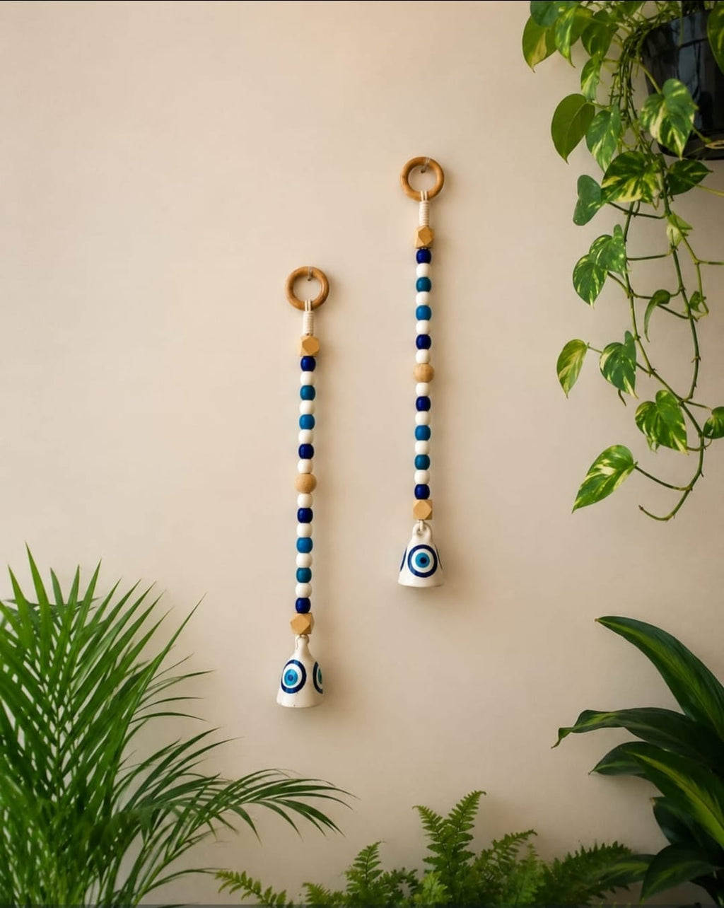 Two hanging beaded wall decorations with wooden rings and evil eye charms, displayed on a beige wall with green plants.