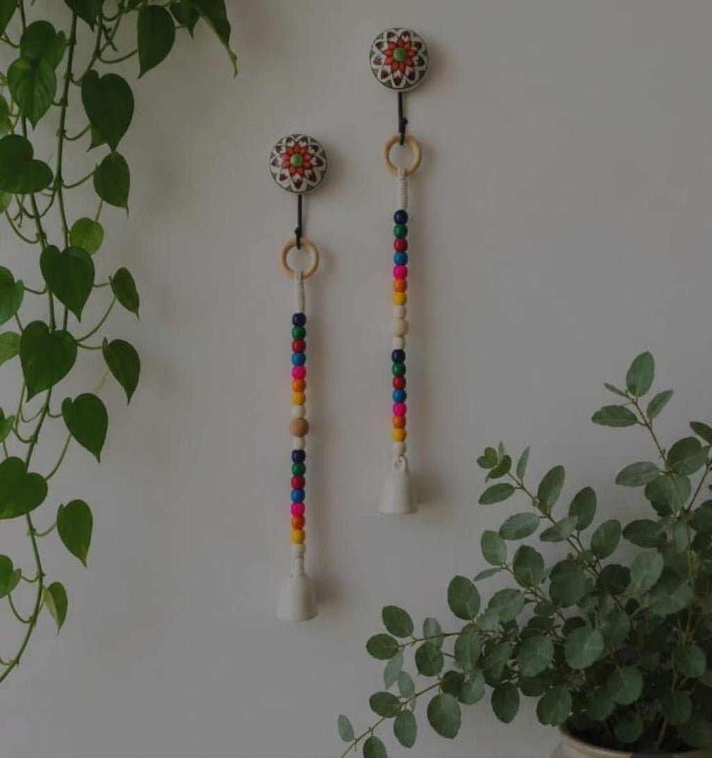 Two bohemian wall hanging ornaments with colorful wooden beads, white bells, and patterned circular tops, displayed on a light wall with green plants.