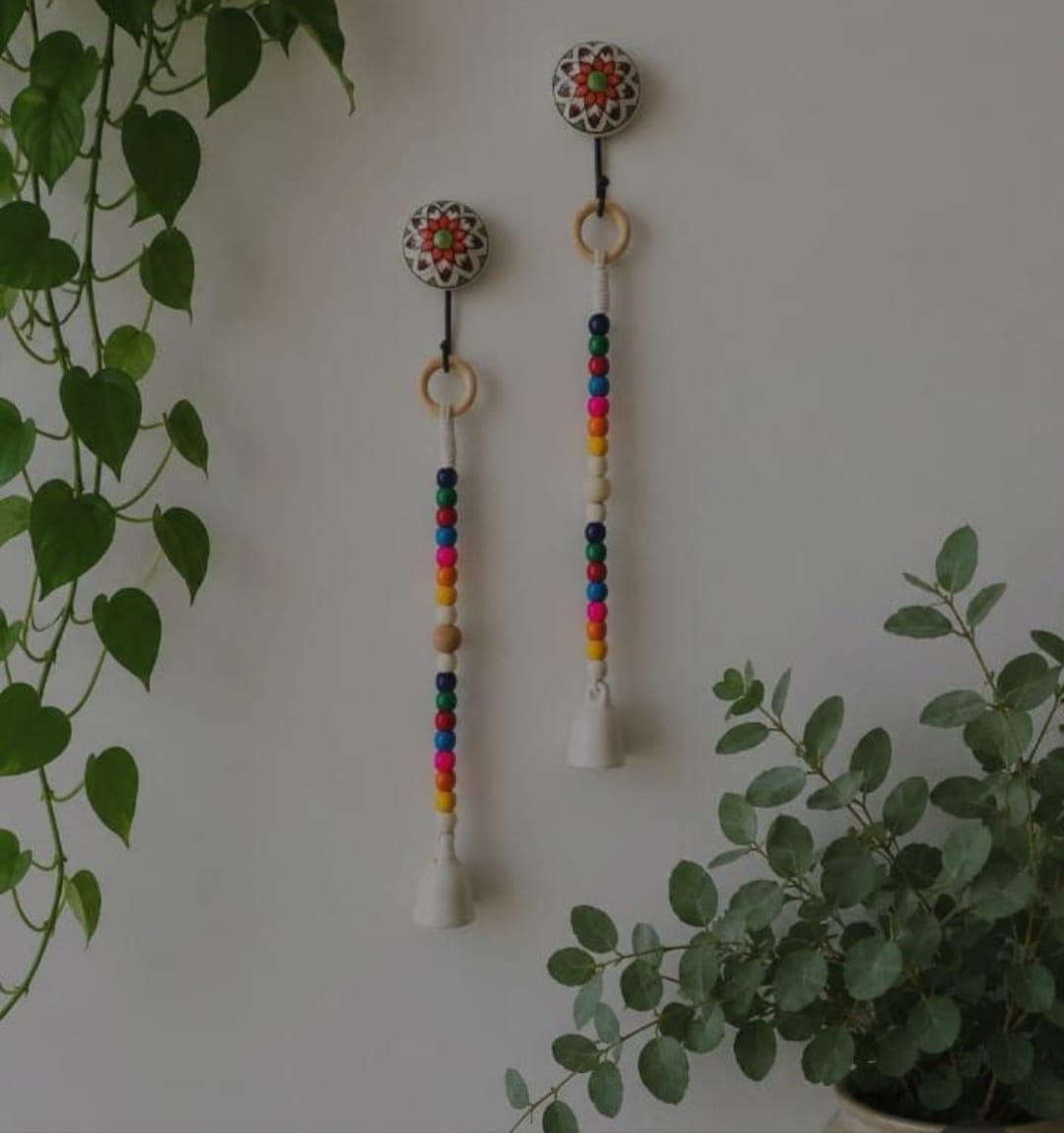 Two bohemian wall hanging ornaments with colorful wooden beads, white bells, and patterned circular tops, displayed on a light wall with green plants.