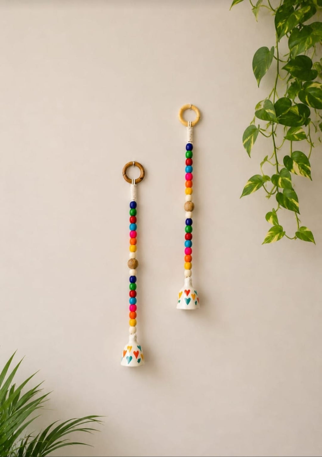 Two colorful beaded wall hanging ornaments with wooden rings and white bell-shaped charms, displayed on a beige wall with green plant leaves.
